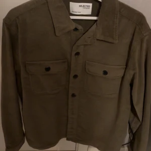 Olivgrön manchesterjacka från Selected - Snygg olivgrön manchesterjacka från Selected med knappar framtill och två bröstfickor. Perfekt för en stilren look. Jackan har långa ärmar och en klassisk krage. Helt oanvänd!