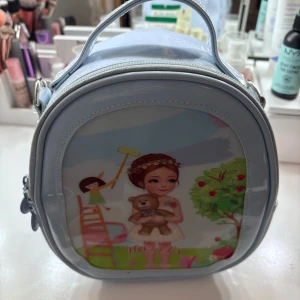 Blå axelväska med illustration - Söt liten blå axelväska med en illustration av en flicka och en nallebjörn på framsidan. Väskan har en dragkedja på baksidan och ett handtag upptill. Perfekt för att bära småsaker med stil. Finns en lång axelband med!