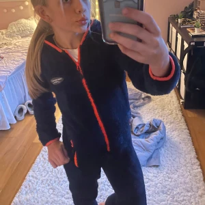 Mörkblå onepiece från Swedemount - Säljer en mysig mörkblå onepicec från Swedemount med orange detaljer. Overallen har en dragkedja framtill och en huva för extra värme. Perfekt för kalla dagar!