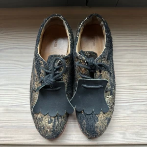 Unika glittriga skor fr. Gram med snörning - Säljer ett par unika skor med glittrigt mönster i svart och guld. Skorna har snörning och en dekorativ detalj framtill. Perfekta för att sticka ut med en cool och annorlunda stil. Från skomärket Gram!  