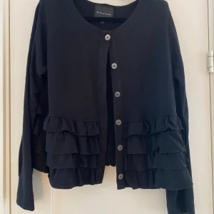 Svart kofta från Bitte Kai Rand strl L - Snygg svart kofta från Bitte Kai Rand med volangdetaljer framtill och knäppning med knappar. Perfekt för att ge en elegant touch till din outfit. Koftan har långa ärmar och en rund halsringning.          Svart kofta med volanger i strl L från Bitte Kai Rand Copenhagen.  Sparsamt använd och i väldigt fint begagnat skick.  Mått Över bröstet: ca 52 cm Längd från axeln och ner: ca 57 cm  53% ull, 43% viskos och 4% polyamid  Kommer från ett djur och rökfritt hem.