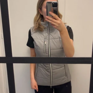 Grå dunväst från Equestrian Stockholm - Snygg grå dunväst från Equestrian Stockholm med dragkedja framtill och två praktiska fickor med dragkedjor. Perfekt att ha i stallet nu när det börjar bli varmare ute 