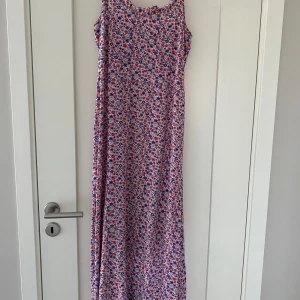 Blommig långklänning - Säljer en vacker blommig långklänning med ett färgglatt mönster i rött, blått och rosa. Klänningen är ärmlös och perfekt för sommarens varma dagar. Den har en luftig passform som ger en skön känsla. Perfekt för en dag på stranden eller en sommarfest.