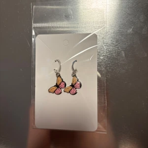 Fjärilsörhängen i rosa och orange - Charmiga örhängen med fjärilsdesign i rosa och orange. Perfekta för att lägga till en färgglad touch till din outfit. De hänger från en silverfärgad krok och är lätta att bära. (Billigt material) 