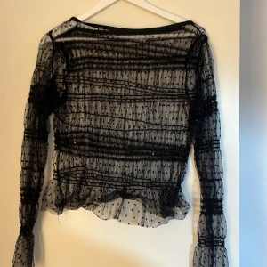 Svart transparent blus med prickar - Säljer en elegant svart transparent blus med prickigt mönster. Blusen har långa ärmar och volangdetaljer vid ärmslut och nederkant. Perfekt för en stilfull look.