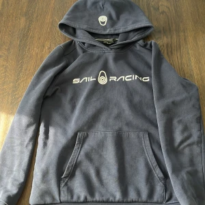 Mörkblå hoodie från Sail Racing - Säljer en snygg mörkblå hoodie från Sail Racing med vit logga på bröstet. Tröjan har en praktisk magficka och en bekväm huva. Perfekt för kyliga dagar!