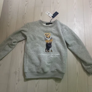 Grå sweatshirt från Polo Ralph Lauren - Säljer en stilren grå sweatshirt från Polo Ralph Lauren med en ikonisk björn på framsidan. Tröjan har långa ärmar och ribbade muddar vid ärmslut och nederkant. Perfekt för en avslappnad look.