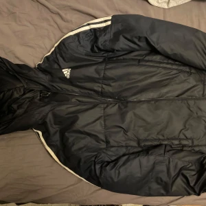 Svart jacka från Adidas - Säljer en svart jacka från Adidas med dragkedja och huva. Jackan har vita detaljer längs ärmarna och är perfekt för kyligare dagar. Adidas-loggan syns tydligt på bröstet. Primegreen-material för en hållbar touch. Herr xs men mer som s.