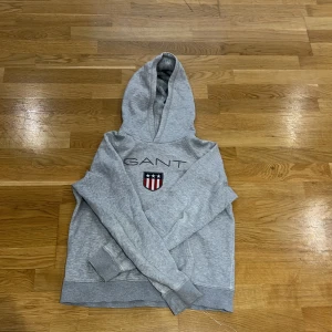 Grå hoodie från GANT - Säljer en stilren grå hoodie från GANT med deras logga och ett emblem på bröstet. Tröjan har en klassisk huva och långa ärmar, perfekt för en avslappnad look.