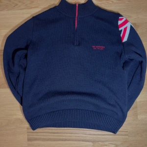 Vintage mörkblå stickad tröja från The Defender - Säljer en mörkblå stickad tröja från The Defender med röd dragkedja och broderad logga. Tröjan har en hög krage med röd insida och ett mönster på axeln i rött och vitt. Perfekt för kyliga dagar!