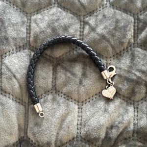 Svart flätat armband med hjärthänge - Snyggt svart flätat armband med ett silverfärgat hjärthänge. Armbandet har en praktisk karbinhake för enkel användning. Perfekt accessoar för att ge en stilren touch till din outfit.