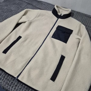 Beige fleecejacka från Livergy - Säljer en beige fleecejacka från Livergy med svarta detaljer. Jackan har en dragkedja framtill och en praktisk bröstficka med lock. Perfekt för kyliga dagar!