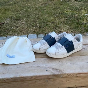 Valentino sneakers med blå detalj - Snygga vita sneakers från Valentino med en bred blå rem över snörningen. Skorna har en klassisk rund tå och en robust sula. Perfekta för en stilren look.