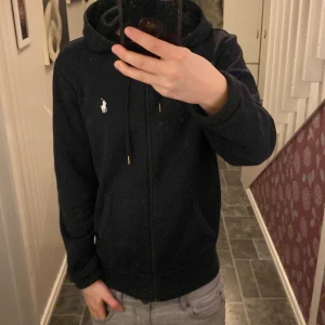 Svart hoodie från Ralph Lauren - Säljer en stilren svart hoodie från Ralph Lauren med dragkedja och klassisk logga på bröstet. Tröjan har långa ärmar och en praktisk huva med snörning. Perfekt för en avslappnad look.