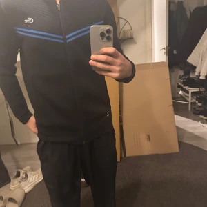 Svart jacka från Lacoste - Säljer en svart Lacoste jacka med blå detaljer. Jackan har dragkedja och långa ärmar. Perfekt för en sportig look med den ikoniska krokodilloggan på bröstet.