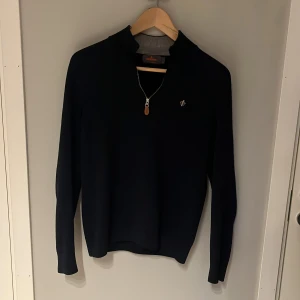 Mörk blå tröja i merinoull från Morris - Snygg svart tröja från Morris i 100% merinoull. Tröjan har en dragkedja vid halsen och en diskret broderad detalj på bröstet. Perfekt för kyligare dagar. Men tyvärr har den ett hål i högra armen som man ser på annonsen.