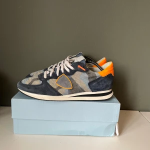 Phillipe models trainers - Snygga sneakers från Philippe Model med ett unikt camouflage-mönster i blå och grå toner. Skorna har orange detaljer och vit snörning. Perfekta för en trendig look. Nypris≈3000kr men mitt pris är 1600kr. Storlek 41. Priset kan sänkas vid snabbare köp. 9/10 i skick. 