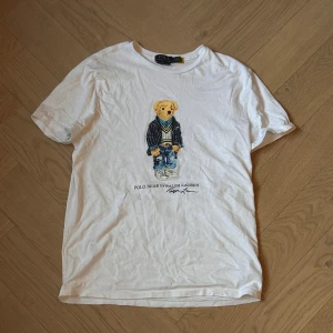 Vit t-shirt med Polo Bear från Ralph Lauren - Säljer en vit t-shirt från Ralph Lauren med det ikoniska Polo Bear-motivet. Björnen är klädd i en stilren outfit med jeans och kavaj. Perfekt för dig som gillar klassisk och lekfull design. T-shirten har en normal passform och är kortärmad.