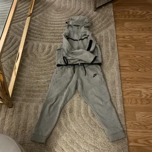 Grå Nike tech fleece sett  - Säljer ett par grå mjukisbyxor från Nike med elastisk midja och snörning. Byxorna har muddar vid bensluten och en liten Nike-logga på benet. Perfekta för en avslappnad stil. Barn storlek 