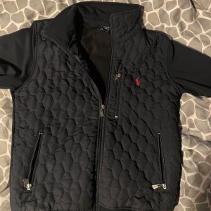 Ralph Lauren cardigan  - Säljer en snygg svart quiltad väst från Ralph Lauren med dragkedja och röd broderad logga på bröstet. Perfekt för lager-på-lager under kyliga dagar. Västen har praktiska fickor med dragkedja. 