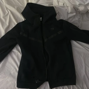 Svart jacka från Nike/ Nike tech - Säljer en stilren svart jacka från Nike med dragkedja och hög krage. Jackan har långa ärmar och en diskret logga på bröstet. Perfekt för en sportig look eller en avslappnad dag.