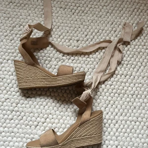 Beige kilklack sandaler från UGG med knytband - Säljer ett par snygga beige sandaler från UGG med hög kilklack och långa band att knyta runt vristen. Ovandelen är i mocka och sulan har flätad jutedetalj för en somrig look. Perfekta till varma dagar och stilrena outfits.