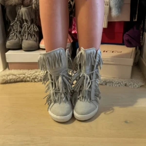 Grå boots med fransar - Säljer ett par grå boots med högt skaft och massor av fransdetaljer. Skorna har snörning framtill och en platt vit sula. Perfekta för dig som vill sticka ut med en unik och trendig stil.