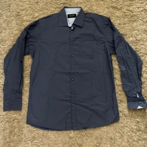 Mörkblå skjorta från Jack Selection - Snygg mörkblå skjorta från Jack Selection med diskret mönster och klassisk krage. Skjortan har långa ärmar och knäppning framtill. Perfekt för dig som vill ha en stilren look.