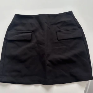 Svart minikjol från Amisu - Säljer en svart minikjol från Amisu med två dekorativa fickor framtill. Kjolen har en enkel och stilren design med dragkedja bak. Perfekt att matcha med olika toppar för en trendig look.