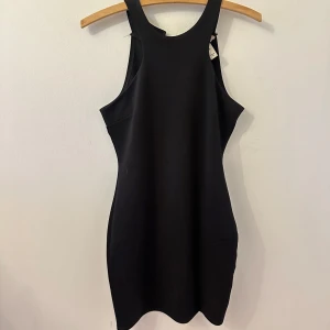 Svart tight ärmlös kortklänning - Säljer en svart tight kortklänning med ärmlös design och hög halsringning. Klänningen har en stilren och minimalistisk look, perfekt för dig som gillar enkla men snygga plagg. Stängs med knappar i nacken.