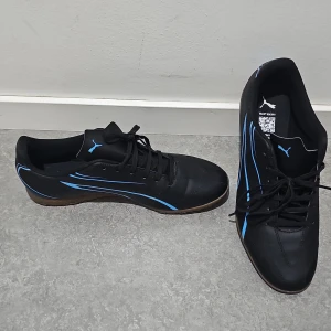 Svarta fotbollsskor från Puma med blå detaljer - Säljer ett par svarta fotbollsskor från Puma med blå detaljer och klassisk snörning. Skorna har rund tå och platt sula, perfekt för dig som gillar att spela fotboll. Puma-loggan syns tydligt på tungan och sidan.