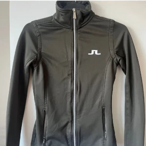 Svart träningsjacka från J.Lindeberg - Sportig svart träningsjacka från J.Lindeberg med hög krage och vit dragkedja. Jackan har två fickor med dragkedja och en vit logga på bröstet. Perfekt för aktiva dagar.