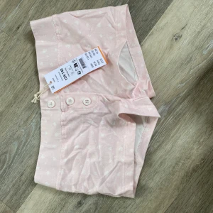 Ljusrosa pyjamasshorts med knappar från ginatricot  - Säljer ett par ljusrosa pyjamasshorts från ginatricot med små vita blommor och tre knappar framtill. Shortsen har en mjuk resår i midjan och är perfekta för mysiga hemmakvällar.