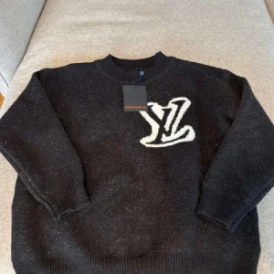 Svart stickad tröja från Louis Vuitton - Säljer en svart stickad tröja från Louis Vuitton med stor vit LV-logga både fram och bak. Tröjan har rund halsringning och långa ärmar med ribbade muddar. Perfekt för dig som gillar exklusiva och stilrena plagg.