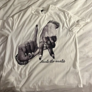 Vit t-shirt med tryck 'Rock the world' - Vit t-shirt med stort svartvitt tryck av två händer med smycken och texten 'Rock the world' på framsidan. Rund halsringning och korta ärmar. Perfekt statement-plagg för dig som gillar coola prints.