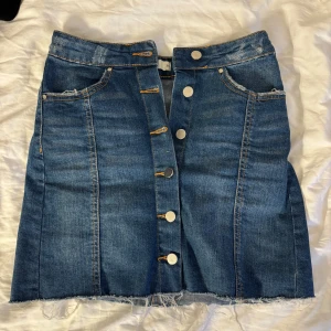 Jeanskjol med knappar framtill - Säljer en klassisk blå jeanskjol med synliga silverfärgade knappar framtill och rå kant nertill. Kjolen har två fickor fram och markerade sömmar som ger en snygg passform. Perfekt till vardags med en croppad tröja eller t-shirt.