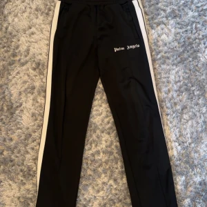 Svarta byxor med vita sidor från Palm Angels - Säljer ett par svarta byxor från Palm Angels med vita ränder längs sidorna och logga tryckt på vänster lår. Byxorna har elastisk midja och raka ben, perfekt för en sportig och trendig look.