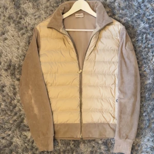 Beige dunjacka från Moncler *2022 årsmodell  - Snygg beige dunjacka från Moncler med quiltad front och ärmar i manchester. Jackan har dragkedja och en klassisk krage. Perfekt för dig som vill ha en stilren och exklusiv look.