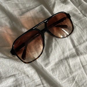 Bruna pilotbågar från Polaroid - Snygga solglasögon från marceti. Bågarna är svarta med bruna tonade glas och har en stilren design. Perfekta för att lyfta din look under soliga dagar.