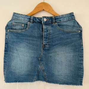 Jeanskjol från Gina  - Säljer en klassisk blå jeanskjol från Gina med rå kant nertill och knappgylf. Kjolen har fyra fickor och en rak passform som slutar ovanför knäna. Perfekt till vardags med en t-shirt eller hoodie.