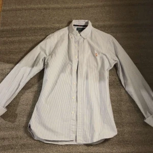 Randig skjorta från Polo Ralph Lauren - Säljer en klassisk blå- och vitrandig skjorta från Polo Ralph Lauren i slim fit. Skjortan har långa ärmar, knappar framtill och broderad logga på bröstet. Perfekt för en stilren look.
