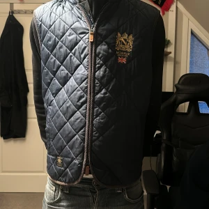 Blå quiltad väst från Morris - Snygg blå quiltad väst från Morris med guldbroderad logga och brittisk flagga på bröstet. Västen har dragkedja framtill och en klassisk, tidlös look. Perfekt för lager-på-lager-stil.