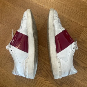 Valentino Rockstud Open sneakers med röd detalj - Säljer ett par vita Valentino Open sneakers med en bred röd lackad rem över mitten. Skorna har snörning och rund tå, samt en platt sula. Klassisk och stilren design med tydlig logga på plösen.