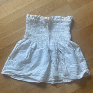Beige sommar kjol från Zara - Säljer en söt vit kjol från Zara med smockad överdel och volangig nederdel. Kjolen har knytband med tofsar framtill och är perfekt för varma dagar.