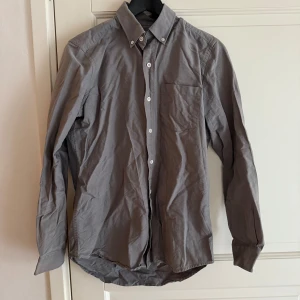 Grå skjorta från A Day's March - Säljer en stilren grå skjorta från A Day's March med klassisk krage och knappstängning framtill. Skjortan har en bröstficka och långa ärmar med manschett. Perfekt till både jeans och chinos. Med en tvätt så är den i toppskick!