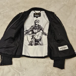 Svart quiltad bomberjacka från Barbour International - Pris kan diskuteras. Säljer en svart quiltad bomberjacka från Barbour International. Jackan har lång ärm, dragkedja och ett unikt innerfoder med motorcykeltryck. Klassisk bombermodell med ribbade muddar och Barbour-logga på ärmen.