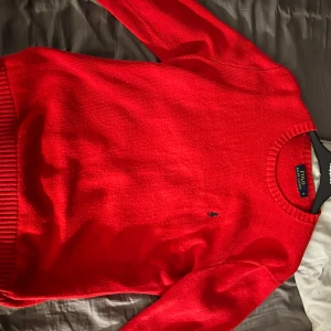 Röd stickad tröja från Polo Ralph Lauren - Klassisk röd stickad tröja från Polo Ralph Lauren med rund halsringning och diskret broderad logga på bröstet. Perfekt för lager-på-lager och passar till många olika stilar.