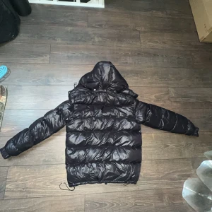Svart dunväst från Moncler - Säljer en svart dunväst från Moncler med hög krage och dragkedja framtill. Västen har en klassisk Moncler-logga på sidan av vänster armen och är perfekt för lager-på-lager-stil. Snygg och varm för kyligare dagar. Storlek monclear: 3