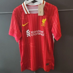 Liverpool hemmaställ 24/25  - Röd Liverpool FC fotbollströja från Nike med tunna vita ränder och gula detaljer. Tröjan har klubbmärke och sponsortryck på bröstet samt vit och gul krage. Perfekt för fans som vill visa sitt stöd på matchdag. Helt ny med prislapp prislapp går att prata om