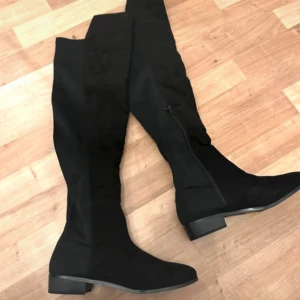 Svarta overknee stövlar med dragkedja - Säljer ett par svarta overknee stövlar med låg klack och dragkedja på insidan. Snygg och stilren modell som passar perfekt till höst och vinter. Gjorda i syntetmaterial med platt sula och rund tå.